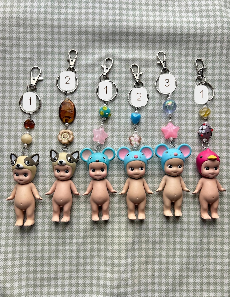 May include: Six keychains with small, nude figurines wearing animal hats. Each keychain has a silver clasp and decorative beads. The hats are designed as a dog, mouse, and bird.
