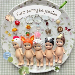 Może przedstawiać: Kolekcja pięciu brelok&oacute;w Sonny Angel z motywem farmy. Każdy brelok przedstawia małą, nagą figurkę w innym kapeluszu zwierzęcym: kurczaka, jelenia, owcy, krowy i niedźwiedzia. Breloki są wyświetlane na ozdobnym talerzu z napisem "farm sonny keychains".