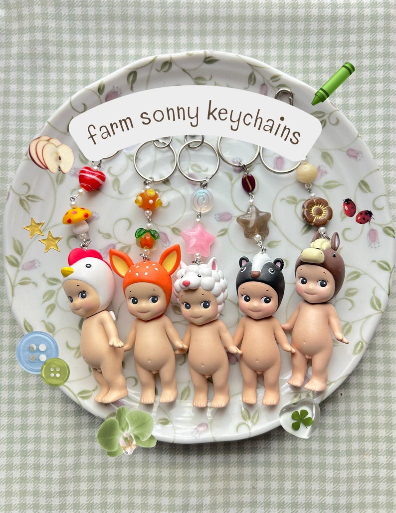 Może przedstawiać: Kolekcja pięciu brelok&oacute;w Sonny Angel z motywem farmy. Każdy brelok przedstawia małą, nagą figurkę w innym kapeluszu zwierzęcym: kurczaka, jelenia, owcy, krowy i niedźwiedzia. Breloki są wyświetlane na ozdobnym talerzu z napisem "farm sonny keychains".