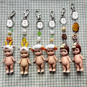 Może przedstawiać: Sześć małych figurek w kolorze sk&oacute;ry z kapeluszami o tematyce zwierzęcej, każda przymocowana do breloczka. Breloki mają ozdobne koraliki i zawieszki. Figurki obejmują kozę, owcę i konia, z numerami 1 i 2.