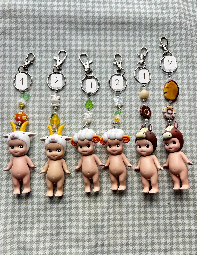 Może przedstawiać: Sześć małych figurek w kolorze sk&oacute;ry z kapeluszami o tematyce zwierzęcej, każda przymocowana do breloczka. Breloki mają ozdobne koraliki i zawieszki. Figurki obejmują kozę, owcę i konia, z numerami 1 i 2.