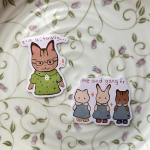 May include: Two stickers featuring cartoon animal characters. One sticker shows a cat wearing glasses and a green shirt with the text "erm actually...". The other sticker shows three animals in blue dresses with the text "me and gang fr".