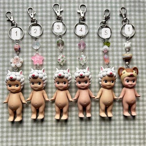 Może przedstawiać: Sześć brelok&oacute;w do kluczy z małymi figurkami w kolorze sk&oacute;ry z kapeluszami zwierząt. Każdy brelok ma srebrne zapięcie, ponumerowany pierścień i ozdobne koraliki w r&oacute;żnych kolorach. Figurki stoją na kratkowanej powierzchni.