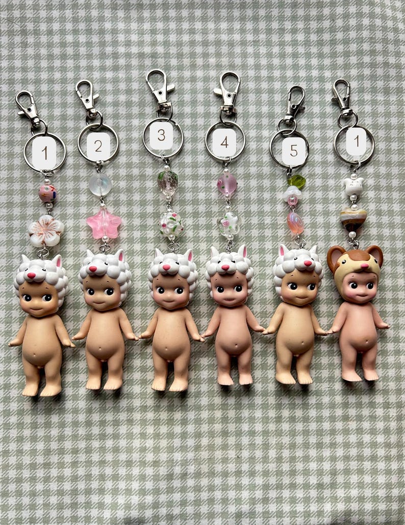 Może przedstawiać: Sześć brelok&oacute;w do kluczy z małymi figurkami w kolorze sk&oacute;ry z kapeluszami zwierząt. Każdy brelok ma srebrne zapięcie, ponumerowany pierścień i ozdobne koraliki w r&oacute;żnych kolorach. Figurki stoją na kratkowanej powierzchni.