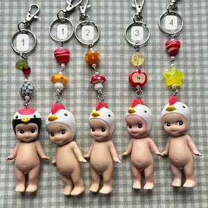 Może przedstawiać: Pięć brelok&oacute;w do kluczy z małymi, nagimi figurkami w kapeluszach z motywem kurczaka. Każdy brelok ma inny urok, w tym grzyb, jabłko i gwiazdę. Breloki mają srebrne okucia.