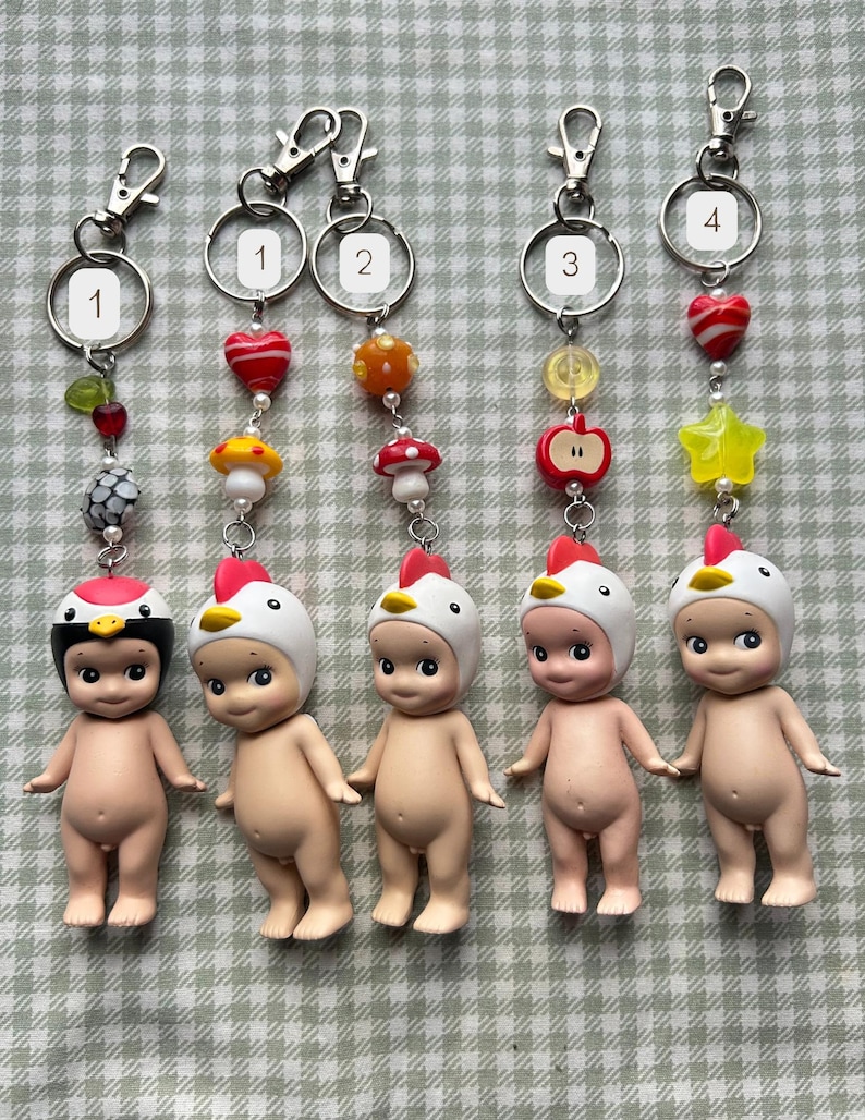 Może przedstawiać: Pięć brelok&oacute;w do kluczy z małymi, nagimi figurkami w kapeluszach z motywem kurczaka. Każdy brelok ma inny urok, w tym grzyb, jabłko i gwiazdę. Breloki mają srebrne okucia.