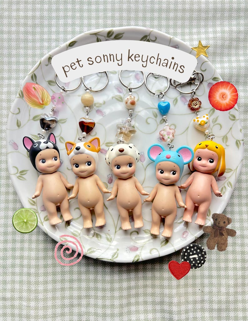 May include: Five Sonny Angel keychains, each with a different animal hat, are arranged on a decorative plate. The plate has the text "pet sonny keychains". The keychains have various charms and beads attached to them.