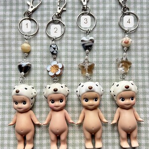 May include: Four keyrings with small, nude baby figurines wearing white hats with black spots. Each keyring has a different design of beads and charms, including hearts, stars, and flowers, attached to a silver ring and clasp.
