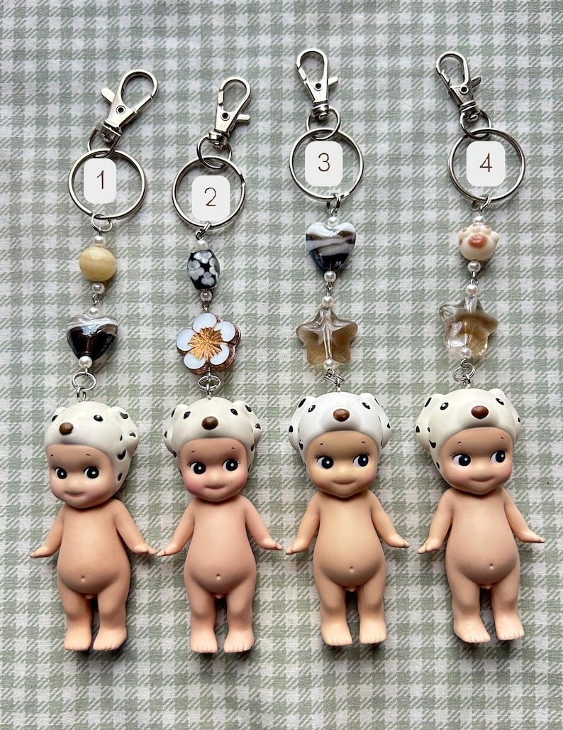 May include: Four keyrings with small, nude baby figurines wearing white hats with black spots. Each keyring has a different design of beads and charms, including hearts, stars, and flowers, attached to a silver ring and clasp.