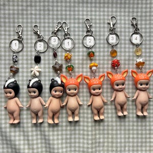 Może przedstawiać: Kolekcja brelok&oacute;w do kluczy z małymi, nagimi figurkami w kapeluszach z motywem zwierzęcym. Każdy brelok ma srebrny zatrzask i ozdobne koraliki. Figurki obejmują wzory skunks&oacute;w i jeleni.