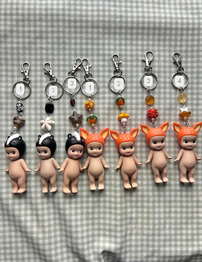 Może przedstawiać: Kolekcja brelok&oacute;w do kluczy z małymi, nagimi figurkami w kapeluszach z motywem zwierzęcym. Każdy brelok ma srebrny zatrzask i ozdobne koraliki. Figurki obejmują wzory skunks&oacute;w i jeleni.