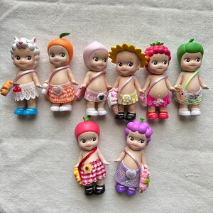 May include: A collection of ten small, colorful dolls with fruit and flower themed outfits. Each doll has a different fruit or flower on their head and a matching outfit. The dolls are all smiling and have big eyes.