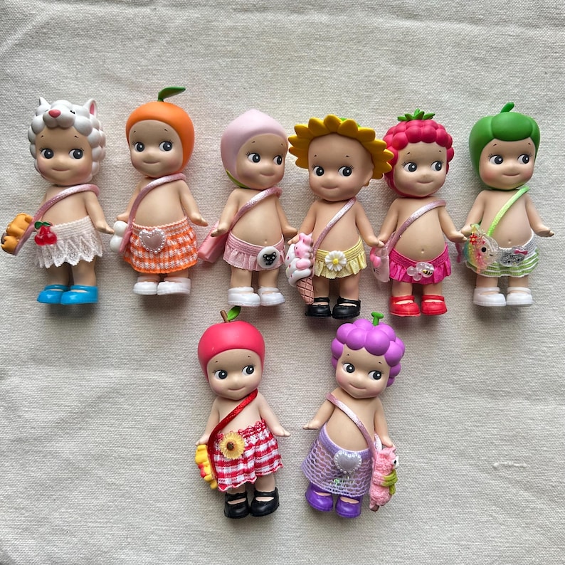 May include: A collection of ten small, colorful dolls with fruit and flower themed outfits. Each doll has a different fruit or flower on their head and a matching outfit. The dolls are all smiling and have big eyes.