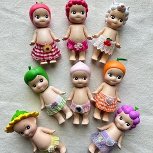 May include: A collection of eight small, colorful dolls with fruit and animal headwear. Each doll is wearing a different patterned skirt or diaper. The dolls are made of plastic and have painted faces.