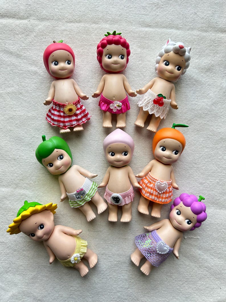 May include: A collection of eight small, colorful dolls with fruit and animal headwear. Each doll is wearing a different patterned skirt or diaper. The dolls are made of plastic and have painted faces.