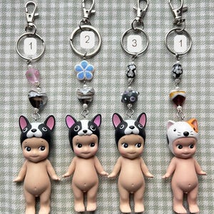 May include: Four keyrings with small figurines. Three figurines wear black and white dog hats, and one wears a white and orange cat hat. Each keyring has a silver ring and a decorative bead chain.