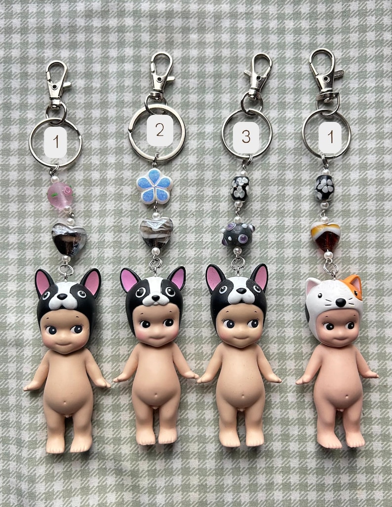 May include: Four keyrings with small figurines. Three figurines wear black and white dog hats, and one wears a white and orange cat hat. Each keyring has a silver ring and a decorative bead chain.