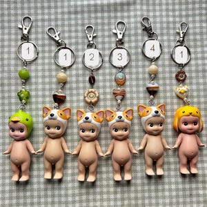 May include: Six keychains featuring small, nude-coloured figurines. Each keychain has a different hat: a duck, a dog, and a yellow-haired figure. The keychains have silver clasps and decorative beads in various colours.