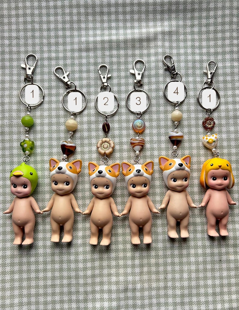 May include: Six keychains featuring small, nude-coloured figurines. Each keychain has a different hat: a duck, a dog, and a yellow-haired figure. The keychains have silver clasps and decorative beads in various colours.