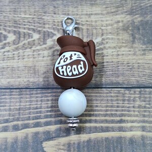 May include: A brown coffee pot charm with a silver lobster clasp at the top. The pot features the white text "Pot Head" on its front. Below the pot is a single round white bead, followed by a small silver decorative spacer bead at the bottom.