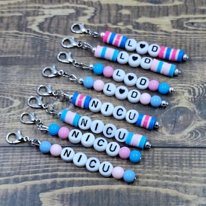 May include: Keychains with white letter beads spelling "LOVE" and "NICU". The keychains have pink, blue, and silver accents. The keychains are on a wooden surface.