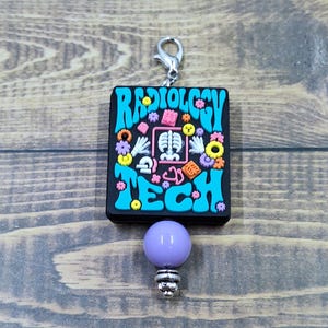 May include: A rectangular black charm featuring the text RADIOLOGY TECH in bright blue letters. The design includes a central pink-framed X-ray of a rib cage, surrounded by small colorful icons of hands, flowers, and gears, with a purple bead below.