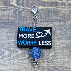May include: A travel-themed badge reel with the text "TRAVEL MORE WORRY LESS" in white and blue. The black rectangular badge has a white airplane and heart graphic. A blue, sparkly, round charm is attached.