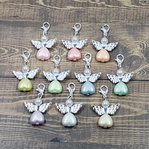 Peut inclure: Une collection de breloques en forme d'ange avec des ailes et des fermoirs argent&eacute;s. Chaque breloque pr&eacute;sente une t&ecirc;te de perle et un corps en forme de c&oelig;ur de diff&eacute;rentes couleurs pastel, dont le rose, le p&ecirc;che, le vert et le bleu. Les breloques sont dispos&eacute;es sur une surface en bois.