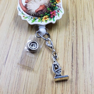May include: A white retractable badge holder with a floral design featuring a mouse. The holder has a silver swivel clip and a silver microscope charm.