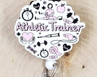 Può includere: Un portabadge bianco e rosa con la scritta "Athletic Trainer" e varie icone mediche e di fitness. Il portabadge ha un cinturino in plastica trasparente e una clip in metallo, su uno sfondo di venature di legno chiaro.