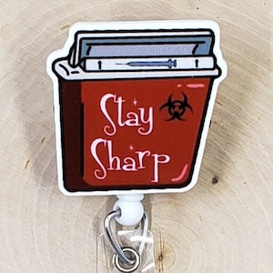 May include: A badge reel featuring a red sharps container graphic with a biohazard symbol and the text Stay Sharp in white script. The container has a blue syringe illustration on the lid. A clear plastic strap with a metal snap button is attached.