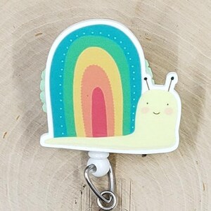 May include: A cute badge reel featuring a smiling cartoon snail with a rainbow shell. The snail is yellow with black antennae and a happy face. The rainbow shell has red, orange, yellow, green, and blue stripes. The reel has a clear plastic strap and a metal clip.