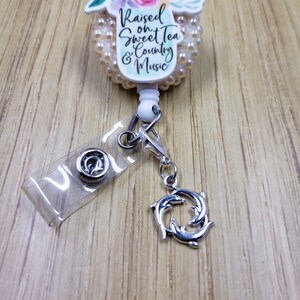 May include: White and pearl-colored badge reel with a floral design and the text "Raised on Sweet Tea & Country Music". The reel is attached to a clear badge holder and a silver dolphin charm.