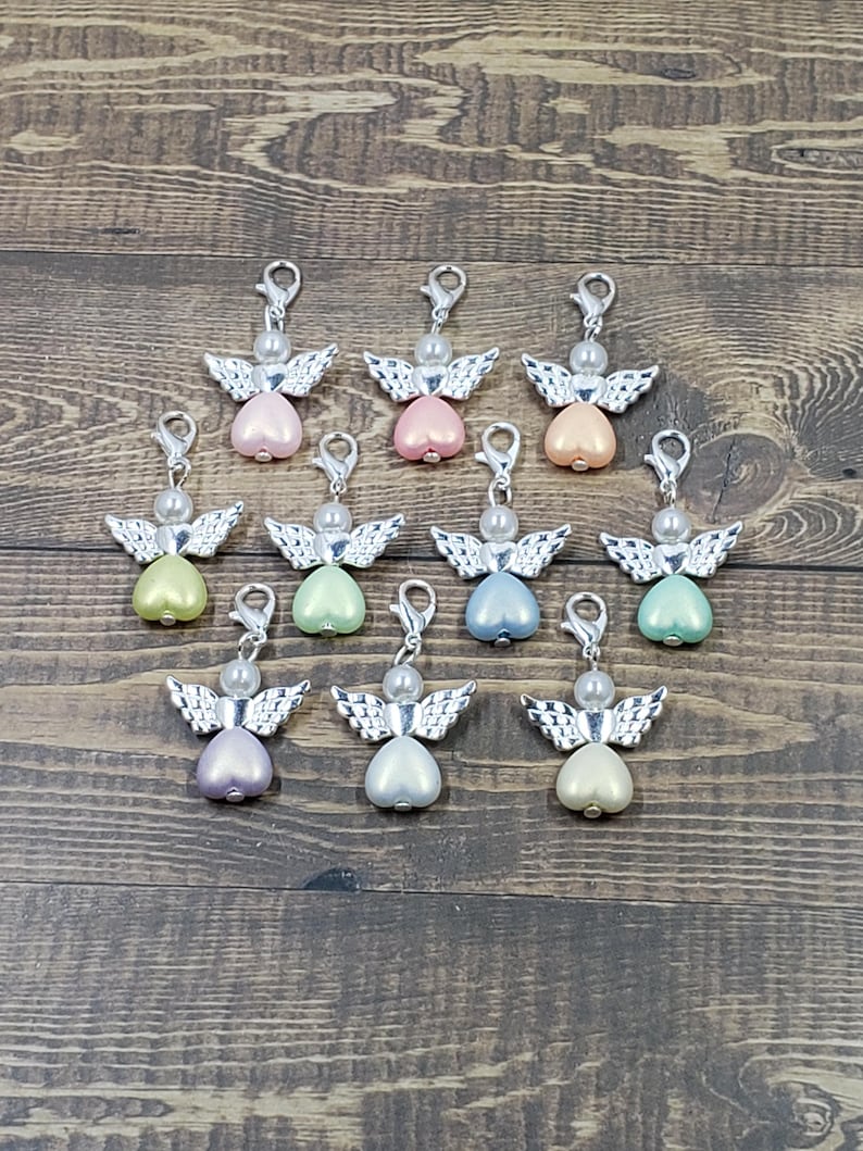 Peut inclure: Une collection de breloques en forme d'ange avec des ailes et des fermoirs argent&eacute;s. Chaque breloque pr&eacute;sente une t&ecirc;te de perle et un corps en forme de c&oelig;ur de diff&eacute;rentes couleurs pastel, dont le rose, le p&ecirc;che, le vert et le bleu. Les breloques sont dispos&eacute;es sur une surface en bois.