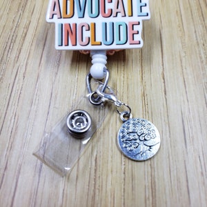 May include: A white retractable badge holder with a colorful "Advocate Include" charm and a silver tree of life charm. The badge holder is on a wooden surface.