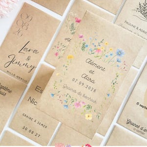 Op de afbeelding: Een verzameling kraftpapieren zaadzakjes met bloemenillustraties en handgeschreven tekst. De zakjes bevatten namen, data en zinnen als "Graines de bonheur" en "Graines d'amour". De ontwerpen omvatten aquarelbloemen in roze, blauw en geel.