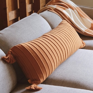 May include: A rectangular lumbar pillow in a solid burnt orange color featuring a ribbed texture and small tassels on each end. The pillow is positioned on a gray sofa alongside a folded throw blanket with a white and burnt orange block pattern.