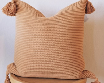 May include: A brown knitted throw pillow with tassels on each corner. The pillow is sitting on a wooden stool.