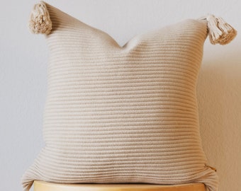 May include: Beige knitted throw pillow with tassel accents. The square pillow features a textured, ribbed design and decorative tassels at each corner. The pillow is placed on a light wooden stool, creating a cozy and inviting aesthetic.