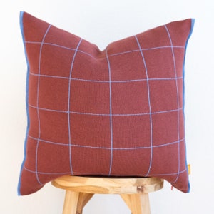 May include: A red pillow with a blue grid pattern and blue trim. The pillow is sitting on a wooden stool.