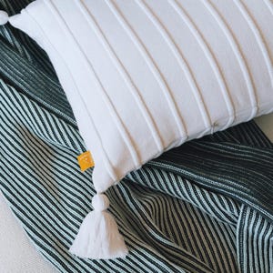 以下が含まれることがあります： 白地にテクスチャ加工が施された枕が、濃い緑と白のストライプの毛布の上に置かれています