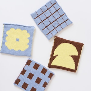 May include: Four square, knitted coasters in various designs. One coaster features a light blue background with a yellow flower design. Another has a light blue background with a brown grid pattern. The other two coasters have brown and light blue patterns.