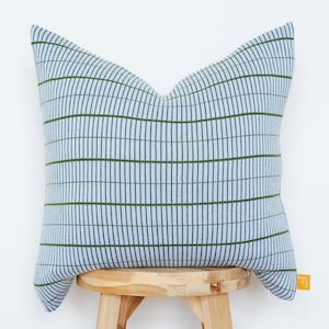 May include: A square decorative pillow with a light blue background and a grid pattern of dark green vertical lines. The pillow is resting on a small wooden stool. The pillow has a tag on the side.