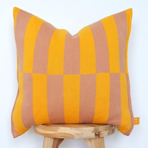 May include: A square cushion with a striking geometric design. It features alternating vertical stripes and squares in shades of golden yellow and a muted peach colour. The cushion is placed on a wooden stool.