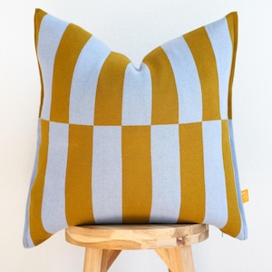 May include: A blue and mustard yellow striped throw pillow with a checkered pattern. The pillow is sitting on a wooden stool.