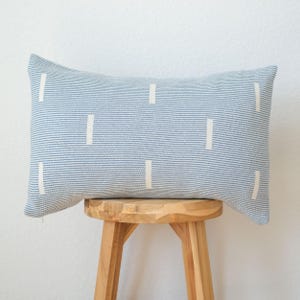 May include: A rectangular decorative pillow with a light blue and white striped pattern. The pillow features vertical white rectangular accents. The pillow is resting on a wooden stool.