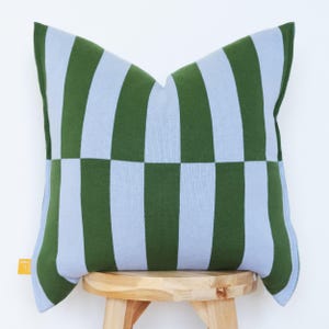 May include: A square decorative pillow with a green and light blue checkerboard pattern. The pillow is made of a soft fabric and is placed on a wooden stool. The pillow has a tag on the side.