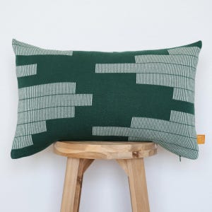 May include: A rectangular throw pillow in a deep forest green color featuring a white geometric pattern of horizontal lines of varying lengths. The pillow has a small orange fabric tag on the right side and a visible zipper along the bottom edge.