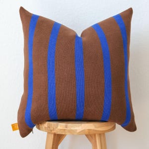 May include: A square throw pillow featuring a brown base with vertical royal blue stripes. The fabric has a visible woven texture. A small orange fabric tag is attached to the lower left corner of the pillow, displaying partial white text.