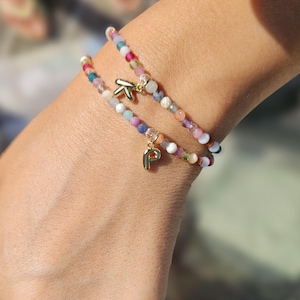 Peut inclure: Deux bracelets de perles avec des pierres précieuses colorées et des breloques dorées. L'un des bracelets porte un charm "K" doré, l'autre un charm "P" doré. Les bracelets sont sur un poignet, avec l'arrière-plan flou.